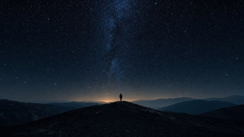 Una persona sola en lo alto de un monte, de espaldas, mirando la Vía Láctea levantarse sobre las montañas