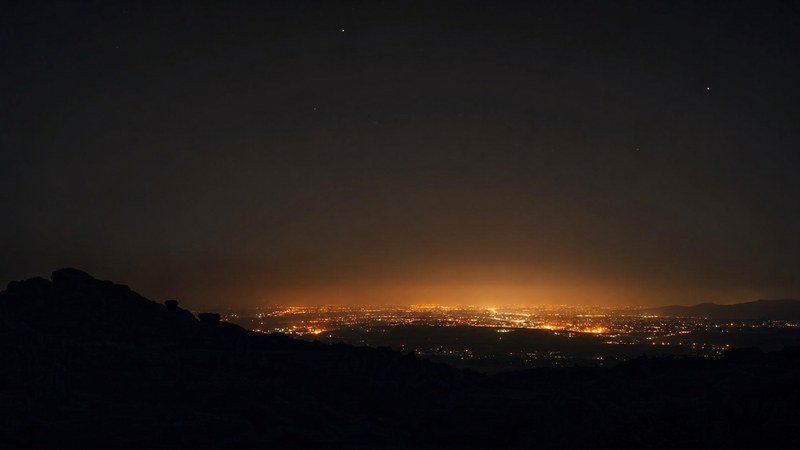 El resplandor naranja de una ciudad visto desde un monte oscuro, con el cielo de encima casi sin estrellas